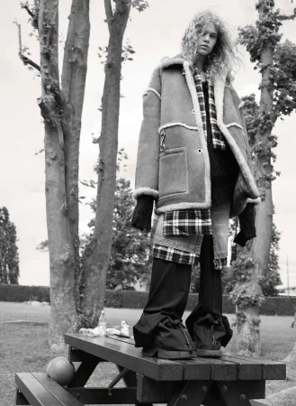 Woman with curly hair stands on a wooden picnic table in a shearling coat, black pants, and gloves in a monochrome outdoor shot.