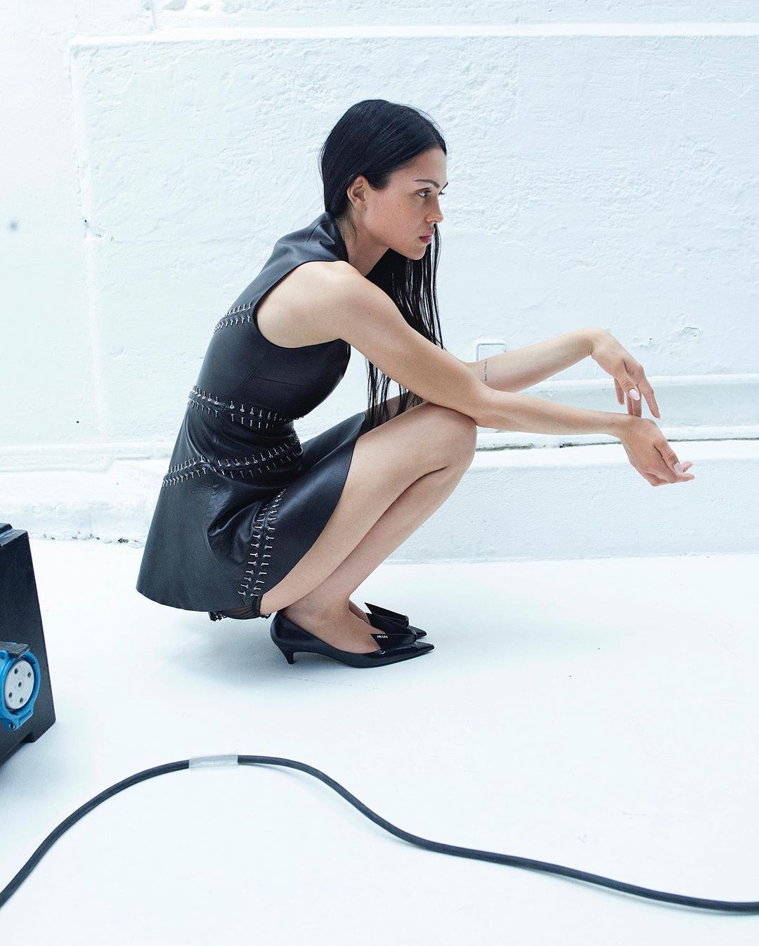 Woman in black dress crouching in a minimalist white photo studio, showcasing fashion and studio photography against a clean background.