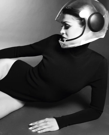 Woman in black bodysuit and clear motorcycle helmet lying on a gray surface in a studio setting, monochrome, high contrast.