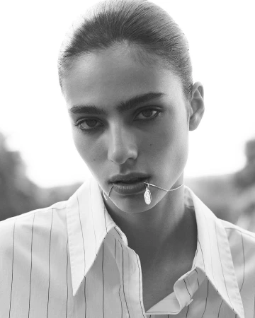 Close up monochrome portrait of a model with hair pulled back wearing a pinstripe button down shirt holding a pendant necklace in her mouth