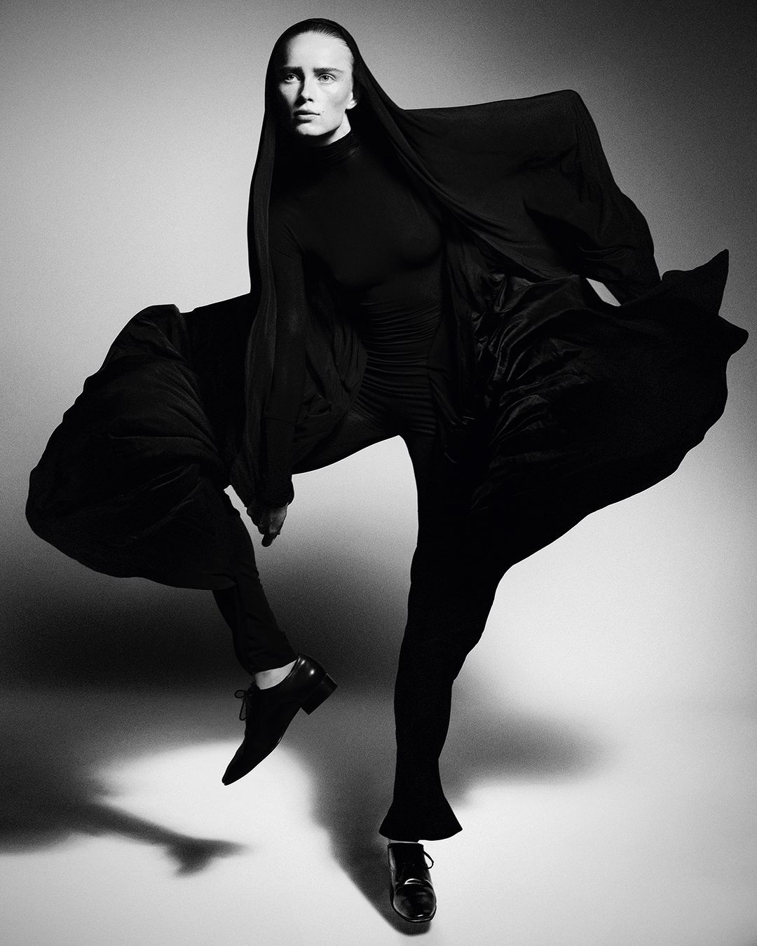Monochrome fashion shot of a single woman in a black turtleneck and flowing black fabric, standing in a dynamic pose on a white background.