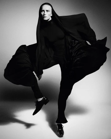 Monochrome fashion shot of a single woman in a black turtleneck and flowing black fabric, standing in a dynamic pose on a white background.