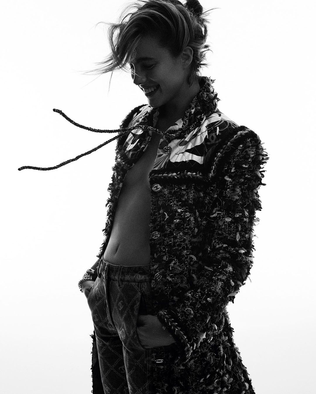 Black and white portrait of a woman wearing a jacket and jeans, smiling with her hands in her pockets, against a white backdrop.