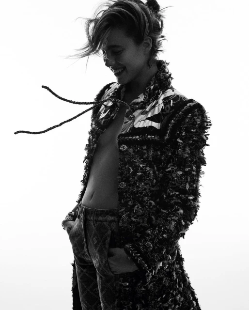 Black and white portrait of a woman wearing a jacket and jeans, smiling with her hands in her pockets, against a white backdrop.