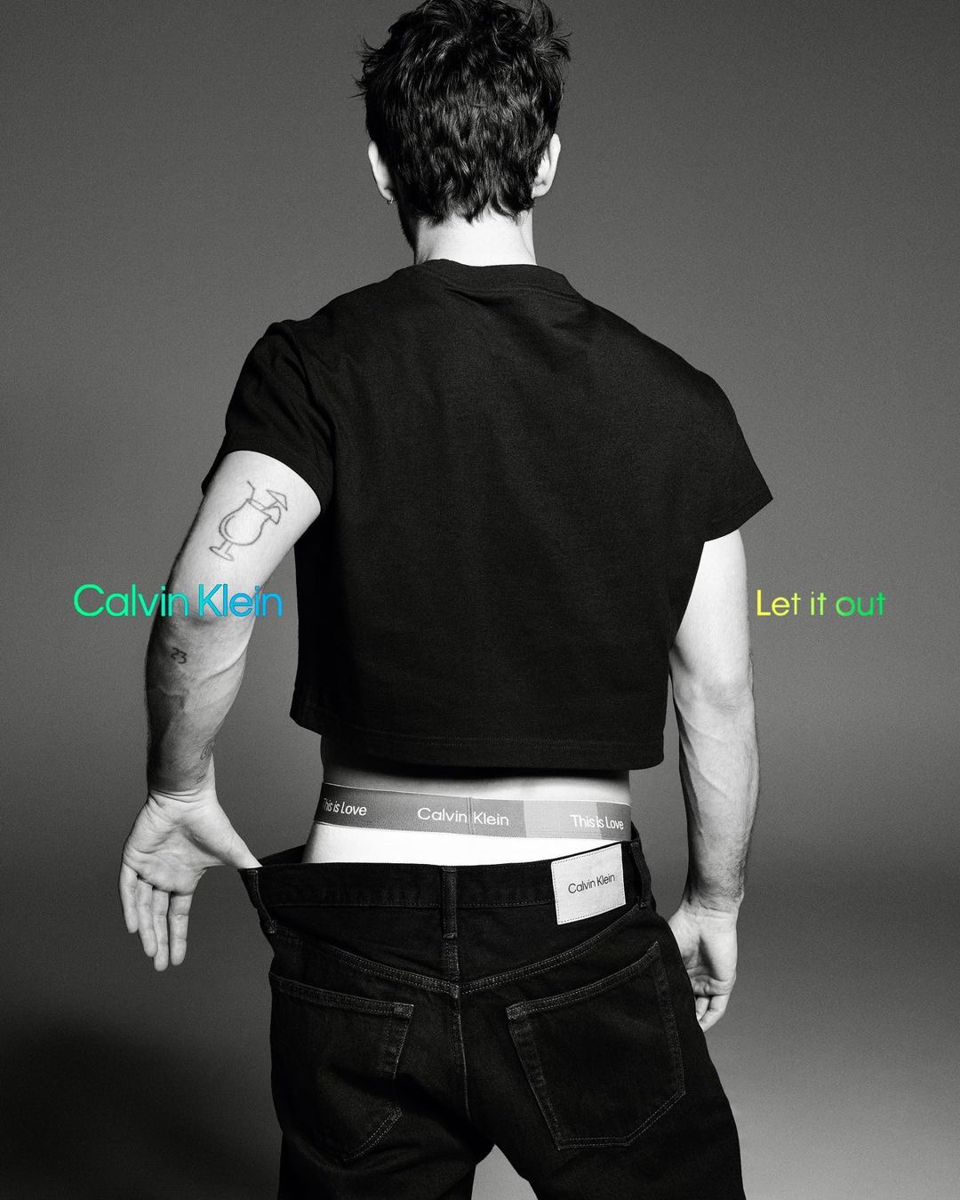 A male model poses from the rear, displaying Calvin Klein underwear, black jeans, and a cropped t-shirt against a grey studio backdrop.