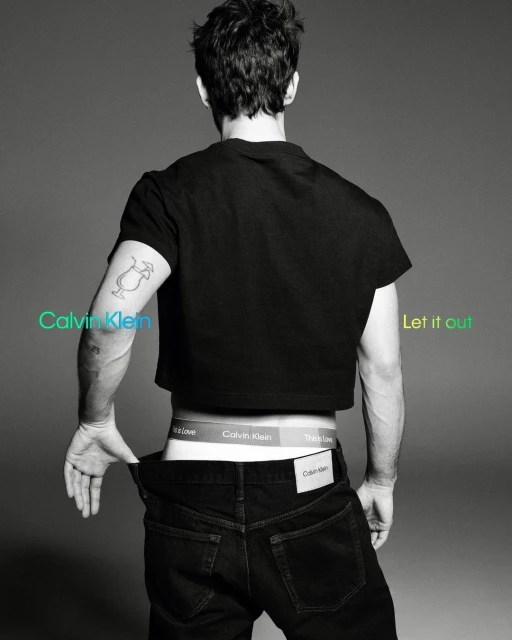 A male model poses from the rear, displaying Calvin Klein underwear, black jeans, and a cropped t-shirt against a grey studio backdrop.