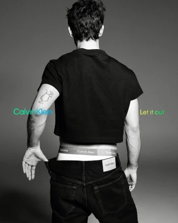 A male model poses from the rear, displaying Calvin Klein underwear, black jeans, and a cropped t-shirt against a grey studio backdrop.