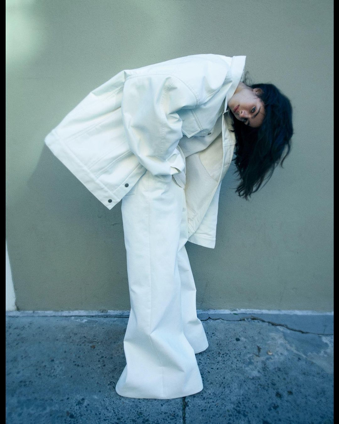 A slender woman with dark hair wearing a white denim jacket and flared pants leans forward in an outdoor urban setting against a wall.