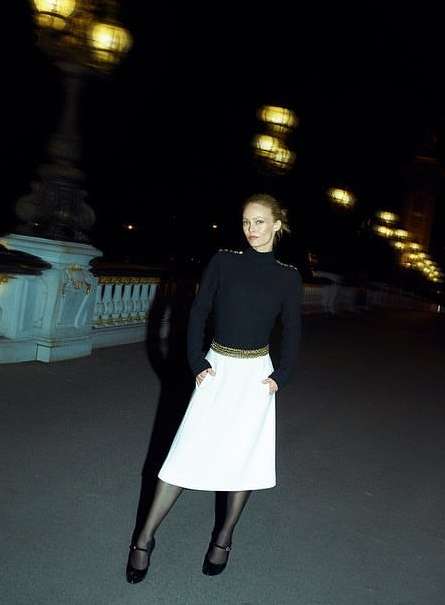 Adult woman poses on a bridge at night with city lights. She wears a black top, white skirt, tights, and Mary Jane shoes.