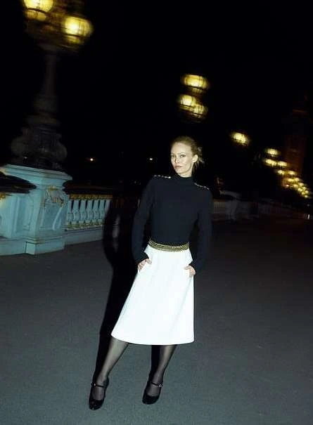 Adult woman poses on a bridge at night with city lights. She wears a black top, white skirt, tights, and Mary Jane shoes.
