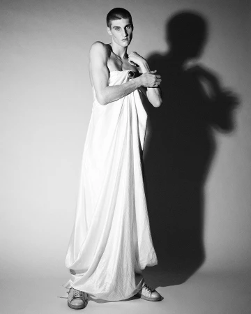 Full-length shot of a fair-skinned male model in white fabric wrap with sneakers, posed against a neutral gradient backdrop with cast shadow.