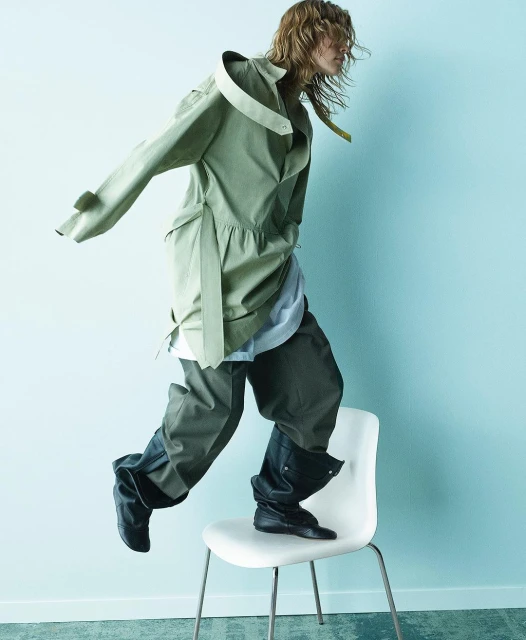 Fashion-forward individual in stylish olive trench coat and dark trousers, captured mid-air on a white chair against a light blue backdrop.