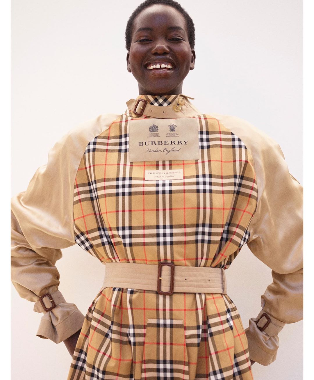 Smiling African woman in a Burberry trench coat with a plaid pattern. Beige fabric and belt. Studio shot with a neutral backdrop.
