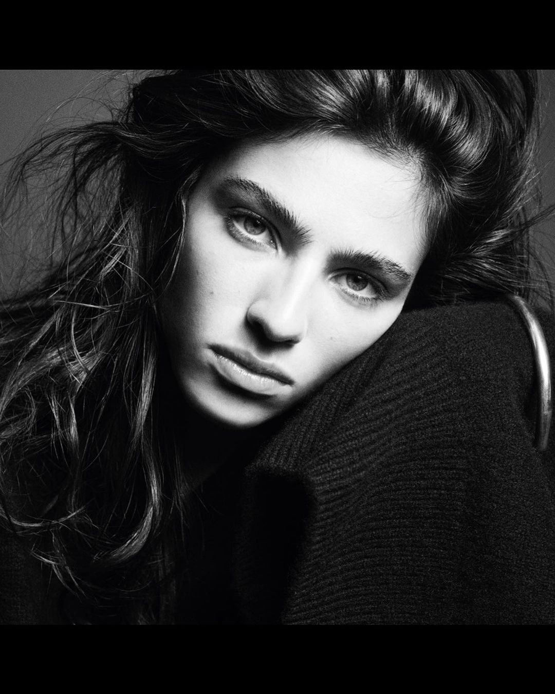 Black and white portrait of a brunette woman with her head resting on her arm, wearing a dark sweater and hoop earring.