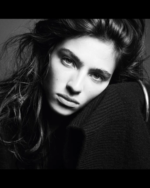Black and white portrait of a brunette woman with her head resting on her arm, wearing a dark sweater and hoop earring.