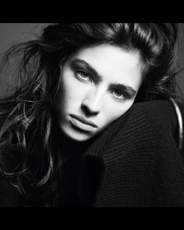 Black and white portrait of a brunette woman with her head resting on her arm, wearing a dark sweater and hoop earring.