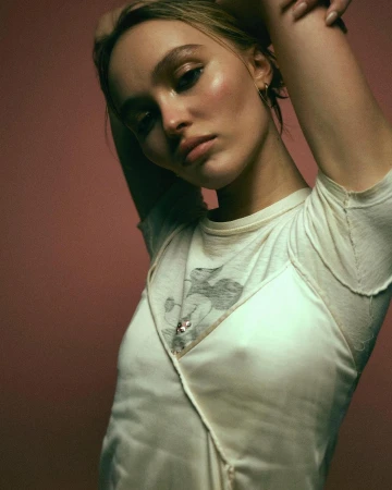 Portrait of Lily Rose Depp wearing an off white layered top with her arms raised against a dark pink backdrop