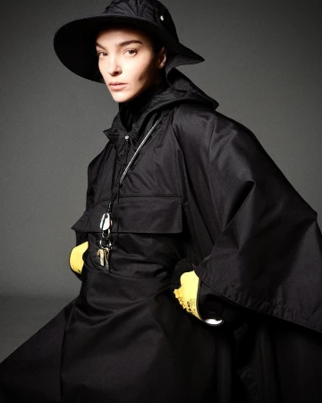 Model wearing a black hooded poncho, matching hat, and yellow gloves against a neutral grey studio background