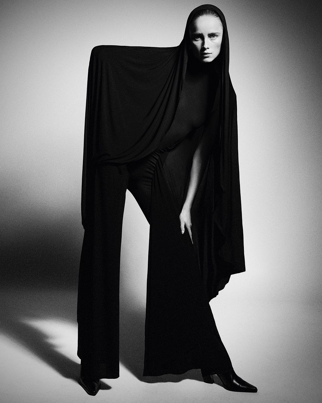 Monochrome studio shot of a tall, slim adult woman posing in elegant black draped clothing with long sleeves and wide-leg pants.