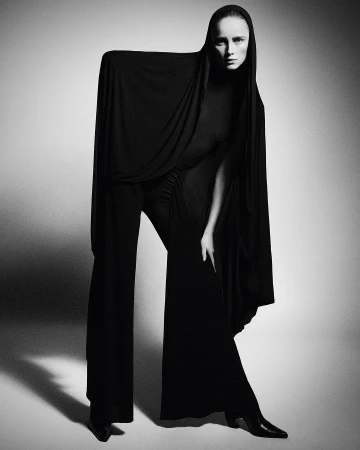 Monochrome studio shot of a tall, slim adult woman posing in elegant black draped clothing with long sleeves and wide-leg pants.