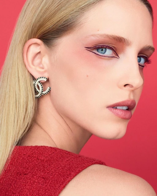 Close-up studio shot of a blonde woman with blue eyes wearing Chanel earrings and vivid graphic eye makeup against a red backdrop.