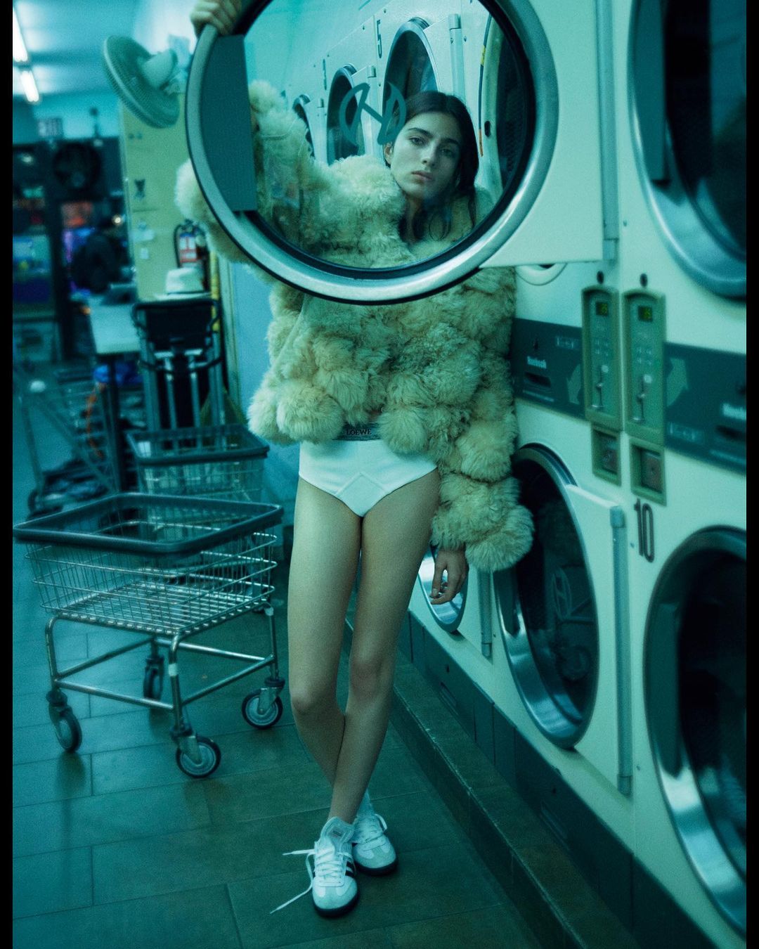 A woman in a fur coat and underwear standing in a laundromat next to washing machines, holding a circular metal frame.