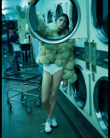 A woman in a fur coat and underwear standing in a laundromat next to washing machines, holding a circular metal frame.