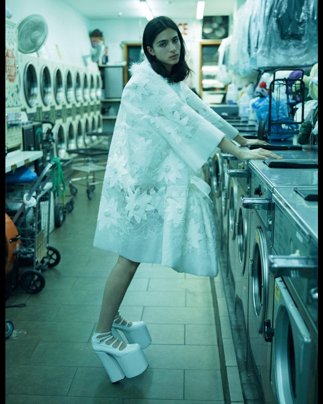 Fashion shot in a laundromat features a woman in a white floral coat and platform shoes, leaning against washing machines.