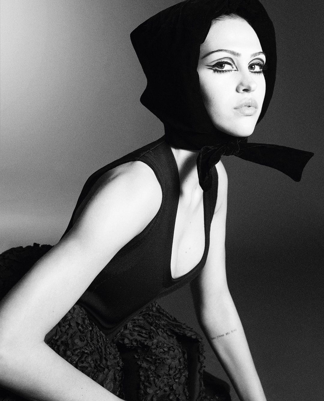 Monochrome portrait of a woman with dramatic eye makeup wearing a black headscarf and a sleeveless top, gazing towards the camera.