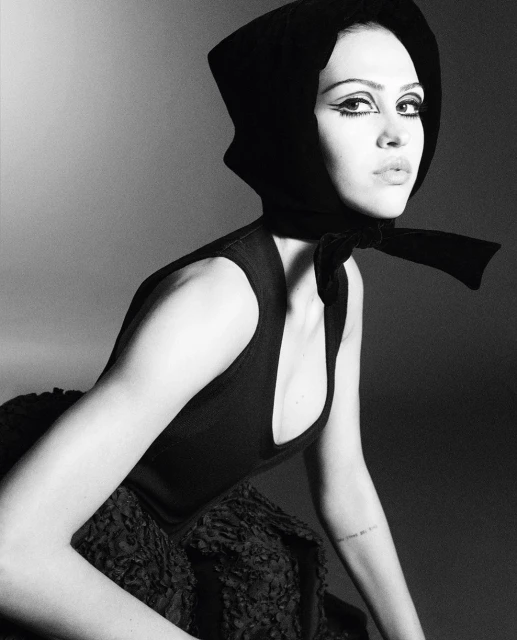 Monochrome portrait of a woman with dramatic eye makeup wearing a black headscarf and a sleeveless top, gazing towards the camera.