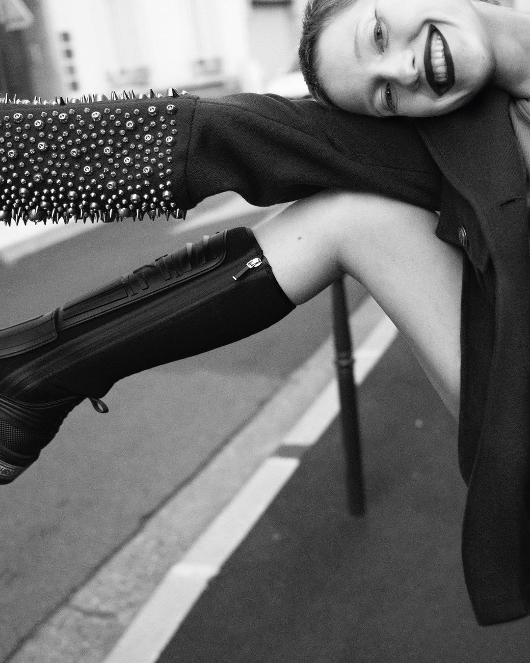 Black and white portrait of a smiling adult female leaning on a studded jacket and shiny black boot, against a neutral urban background.