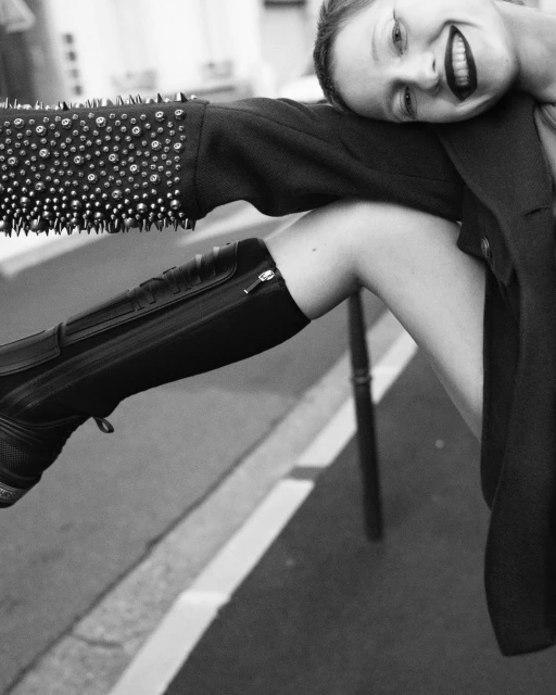 Black and white portrait of a smiling adult female leaning on a studded jacket and shiny black boot, against a neutral urban background.