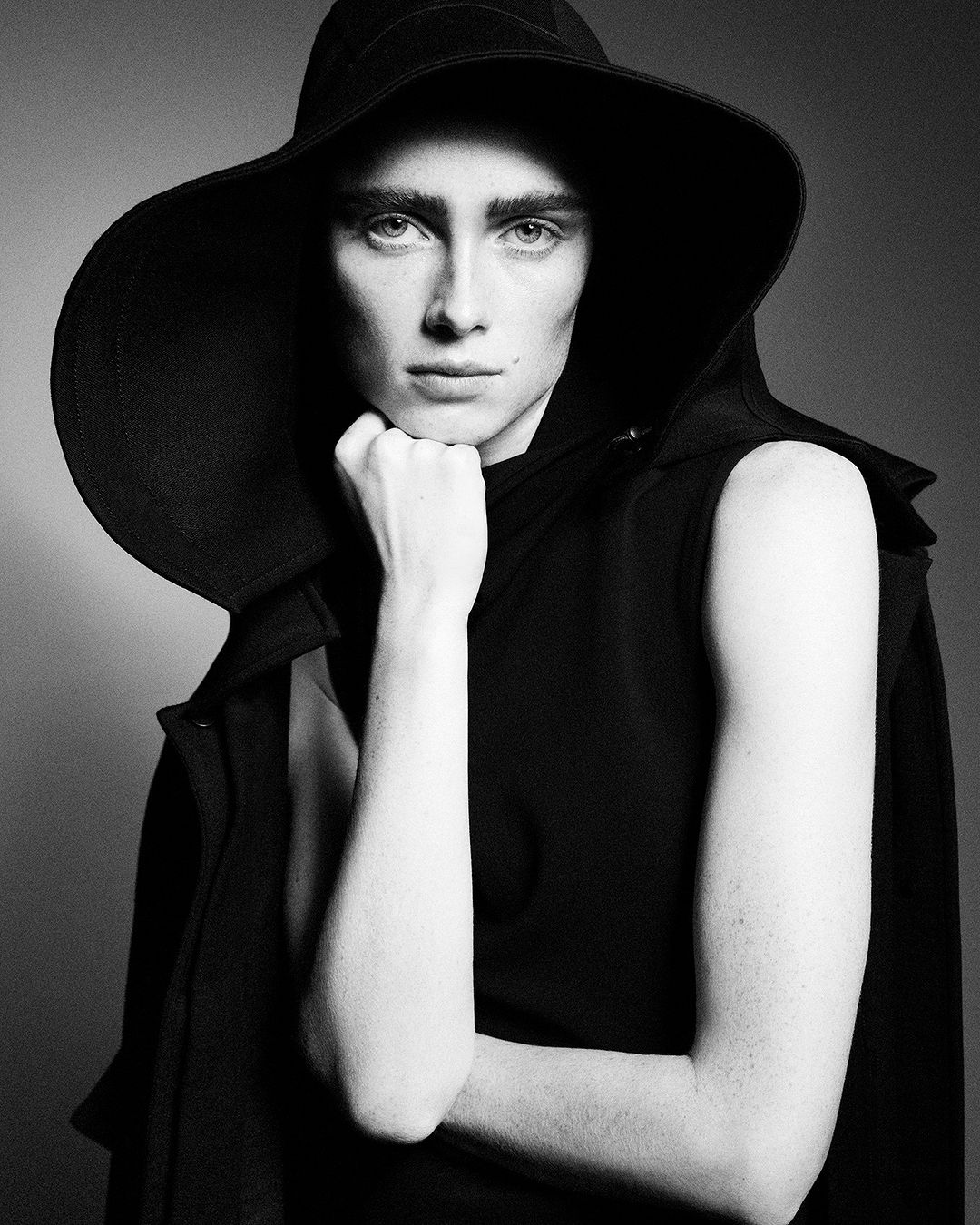 Black and white portrait of a young adult with light skin and eyes, wearing a dramatic wide-brimmed hat and sleeveless coat. 
