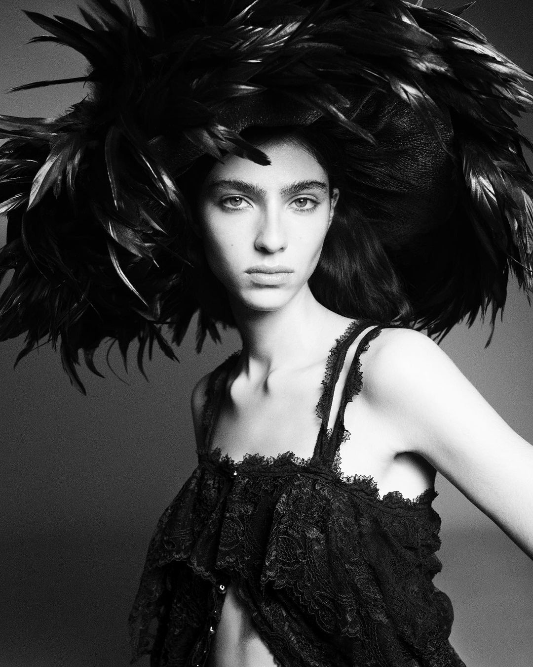 Monochrome portrait of a woman wearing a large feathered hat and lace top, looking directly at the camera in a studio setting.