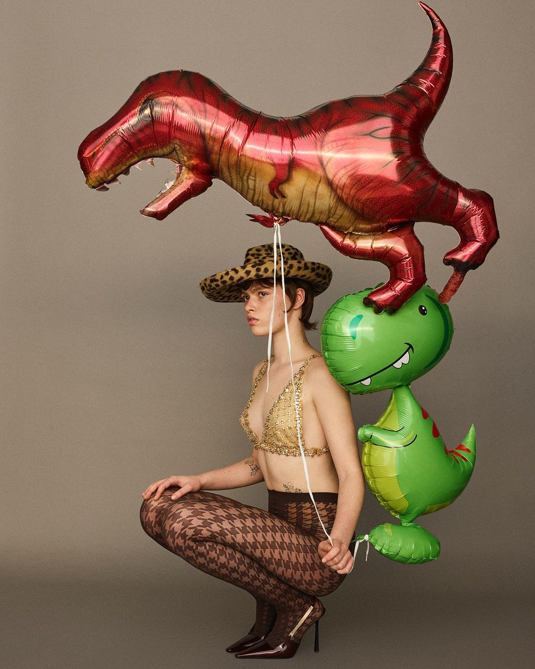 Woman in a hat and bra top poses while holding dinosaur balloons in a studio setting, fashion photography. 