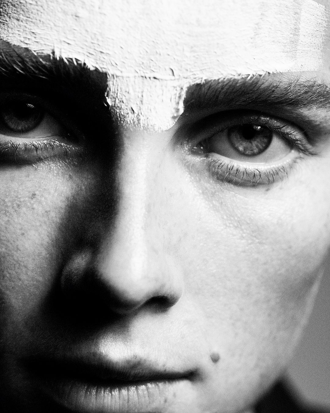 Close-up portrait of a fair-skinned young adult with green eyes and white paint on forehead, captured in black and white.