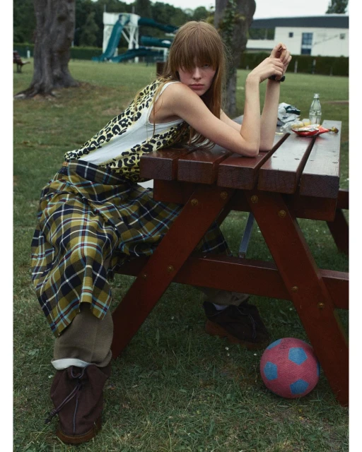 Woman with bangs at picnic table in grassy park setting. Leopard print top, plaid skirt, brown boots, soccer ball, trees, water slide.