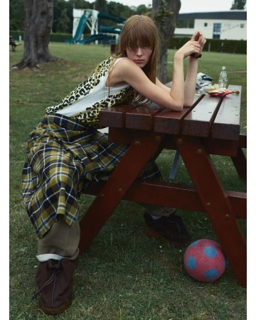 Woman with bangs at picnic table in grassy park setting. Leopard print top, plaid skirt, brown boots, soccer ball, trees, water slide.