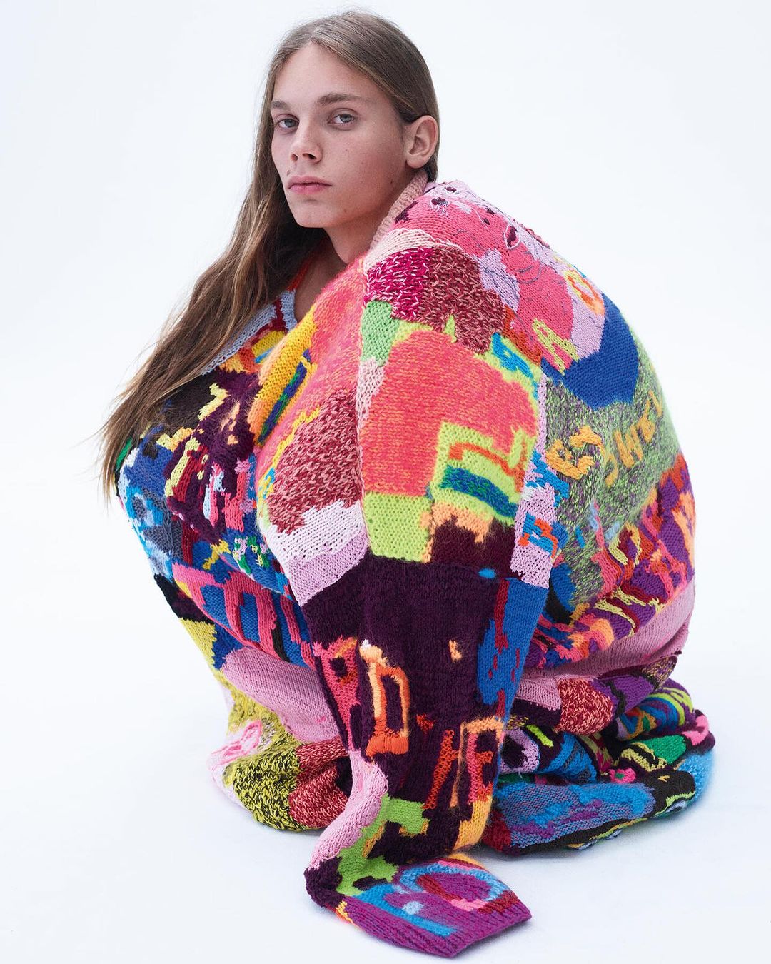 A fashion shot of a person with long brown hair, kneeling while wearing a colorful, patterned sweater against a white background.