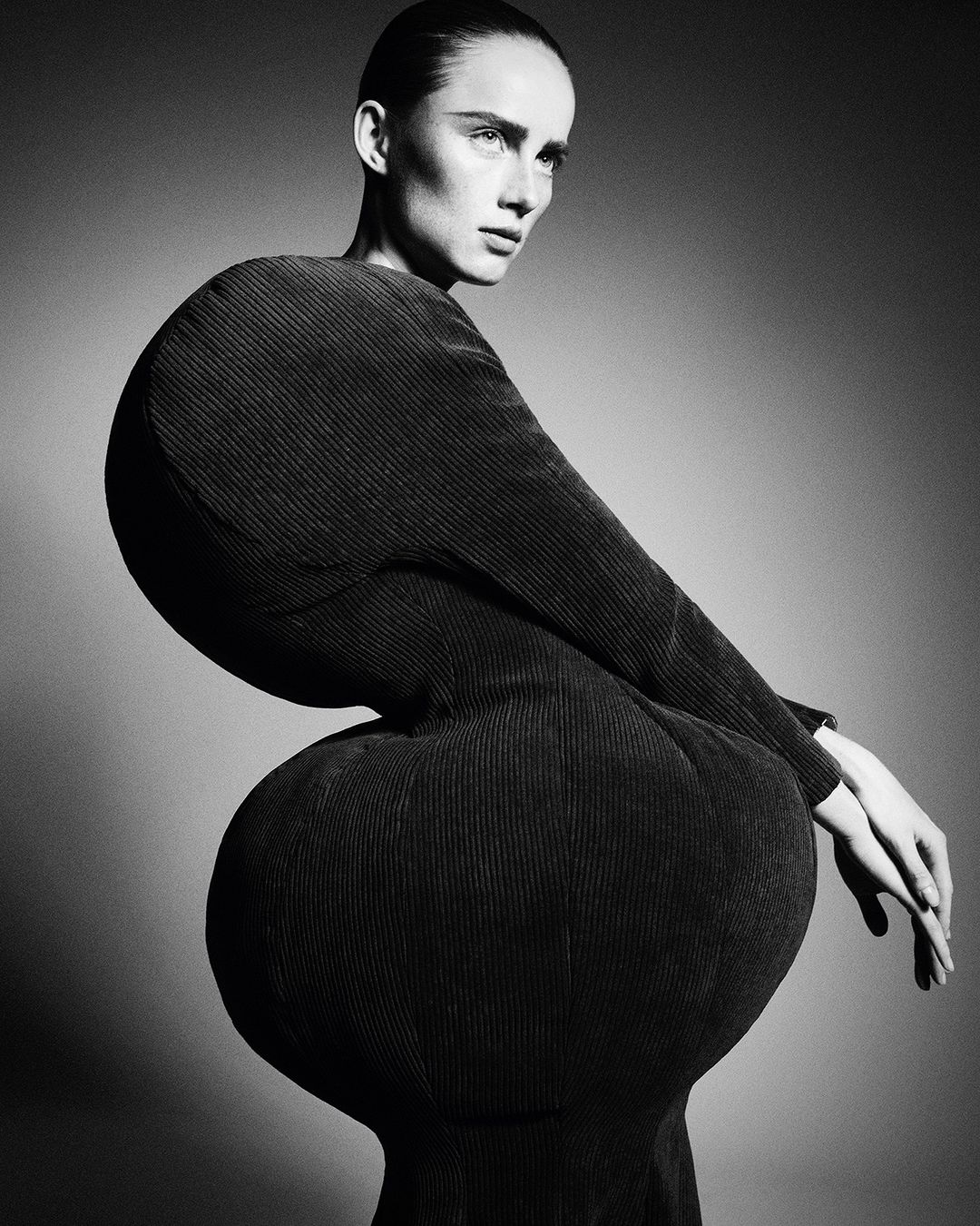 Black and white portrait of a woman in a sculptural black dress, shot in a studio. The dress has rounded shapes at the shoulders and hips.