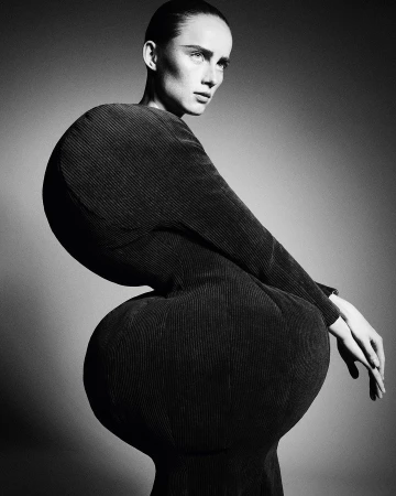 Black and white portrait of a woman in a sculptural black dress, shot in a studio. The dress has rounded shapes at the shoulders and hips.