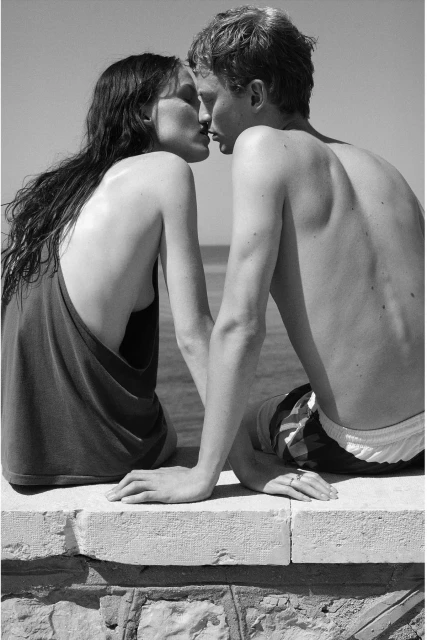 Black and white portrait of a couple kissing on a stone wall near the ocean on a sunny day, with clear sky and ocean water backdrop.