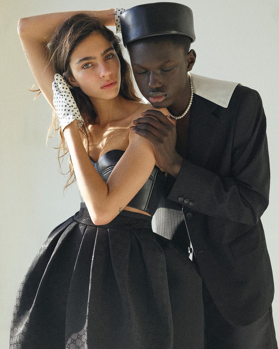 A stylish studio shot captures a young couple, woman in black corset and skirt, man in suit with pearl necklace, posing in soft, neutral light.