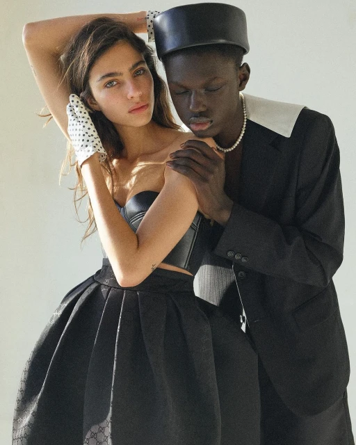 A stylish studio shot captures a young couple, woman in black corset and skirt, man in suit with pearl necklace, posing in soft, neutral light.