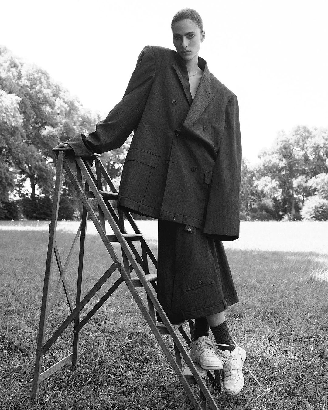 Woman in oversized pinstripe suit poses on a metal ladder outdoors in a monochrome fashion editorial. Stylish outerwear and footwear.