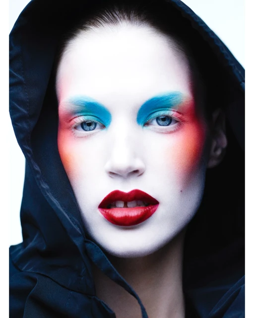 Close-up portrait of a woman with fair skin and colorful makeup, including red lipstick, blue eyeshadow, wearing a hooded jacket.