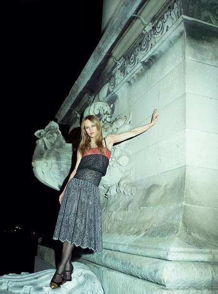 Woman in dress poses near building with architectural details at night. She wears a midi skirt, hose, and high heels.