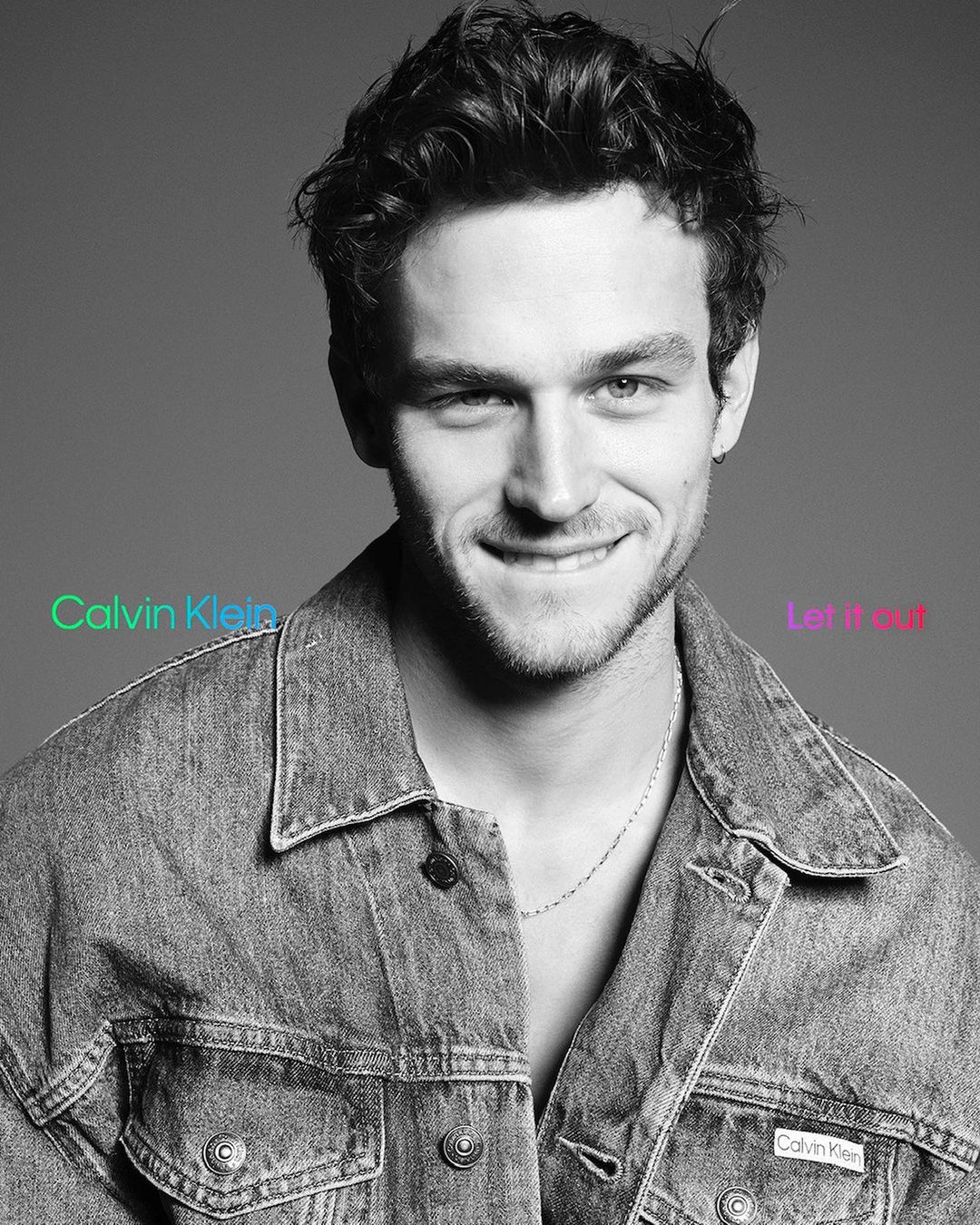 Monochrome portrait of a smiling young man in a denim jacket against a neutral grey background.