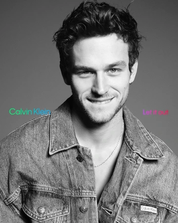 Monochrome portrait of a smiling young man in a denim jacket against a neutral grey background.
