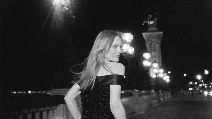 Black and white night photograph of a woman with long hair looking over her shoulder on an illuminated bridge.